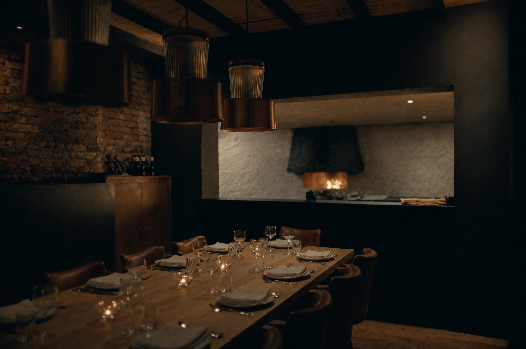 The private room: a long oak table set for fourteen under copper lamps, with the hearth glowing through a window in the far wall.