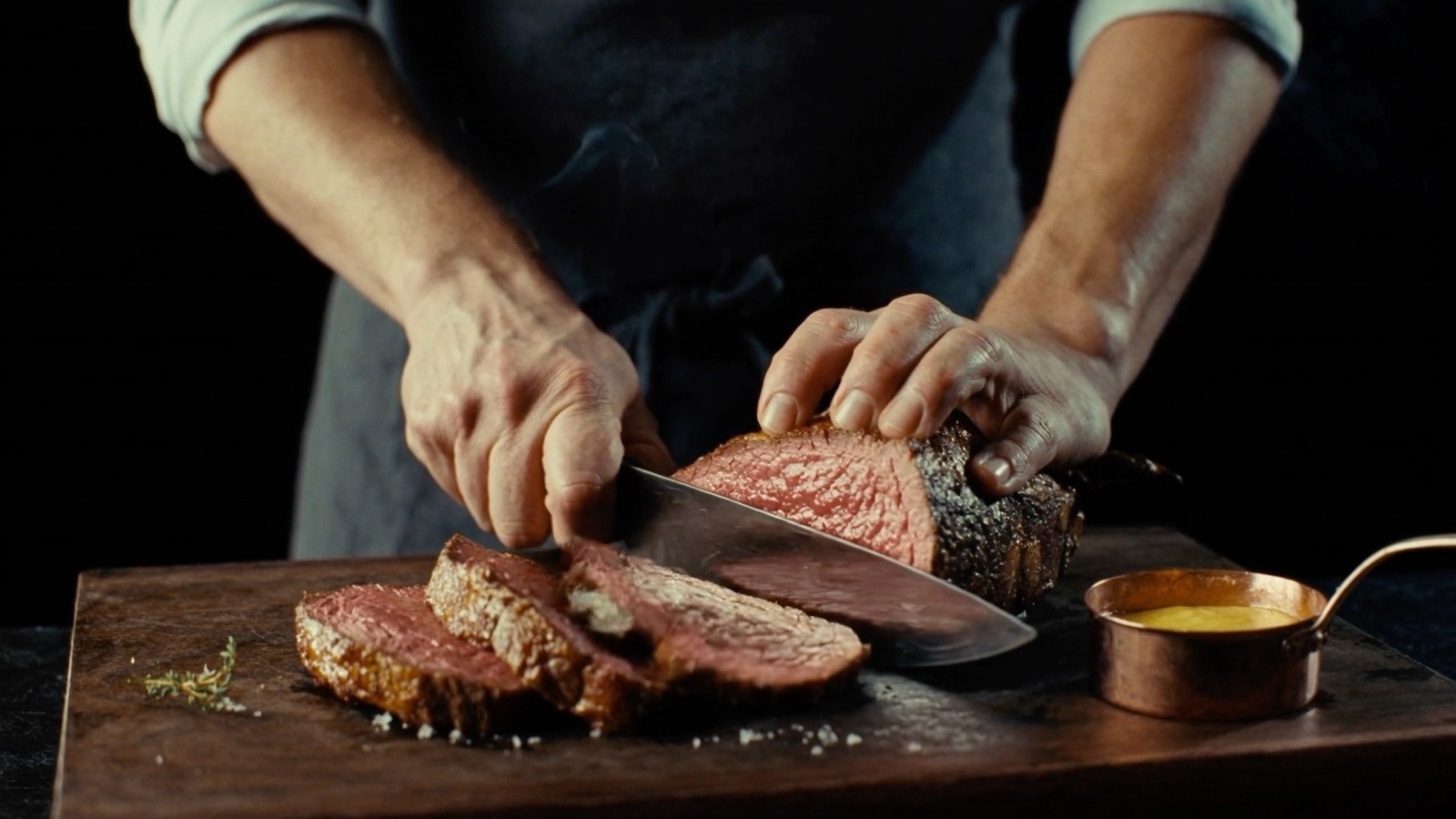A cook slicing a dry-aged strip steak on a wooden board, with béarnaise in a copper pot.