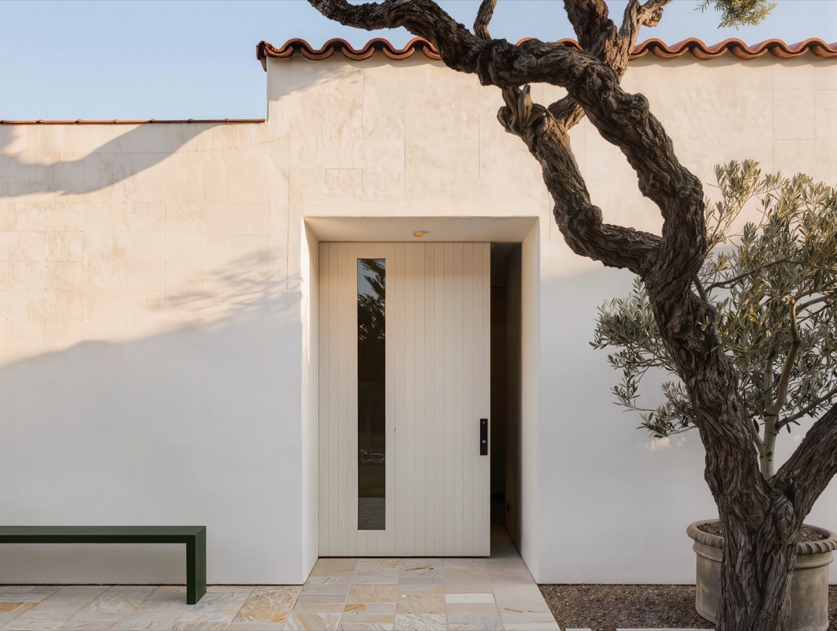 The front entrance: a white plaster wall under a clay tile roof, a pale oak door with a narrow glass panel, a green bench and an old olive tree in the morning sun