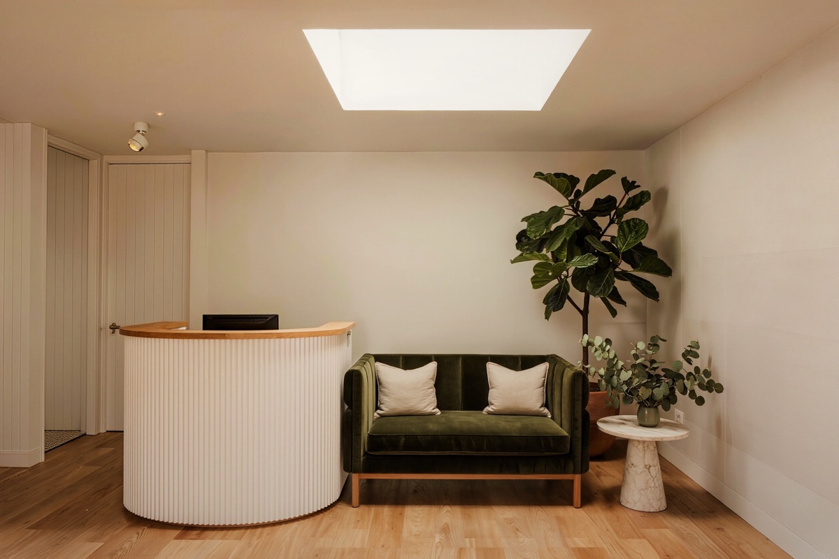 The front room: a curved white oak desk with a fluted front, a green velvet bench with linen cushions, a fiddle-leaf fig and a stone side table with eucalyptus, under a square skylight