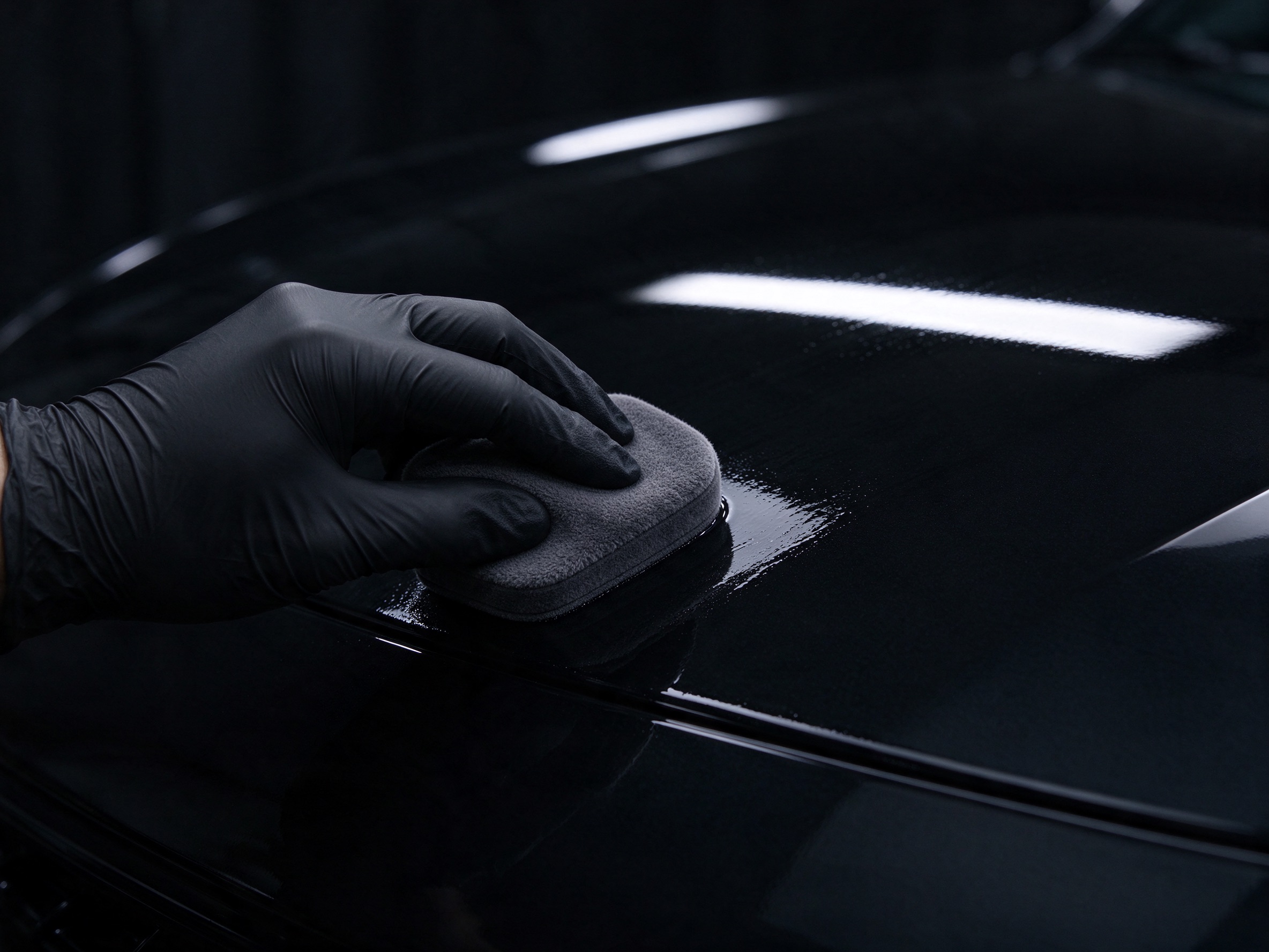 A gloved hand laying ceramic coating on a black panel with an applicator pad.